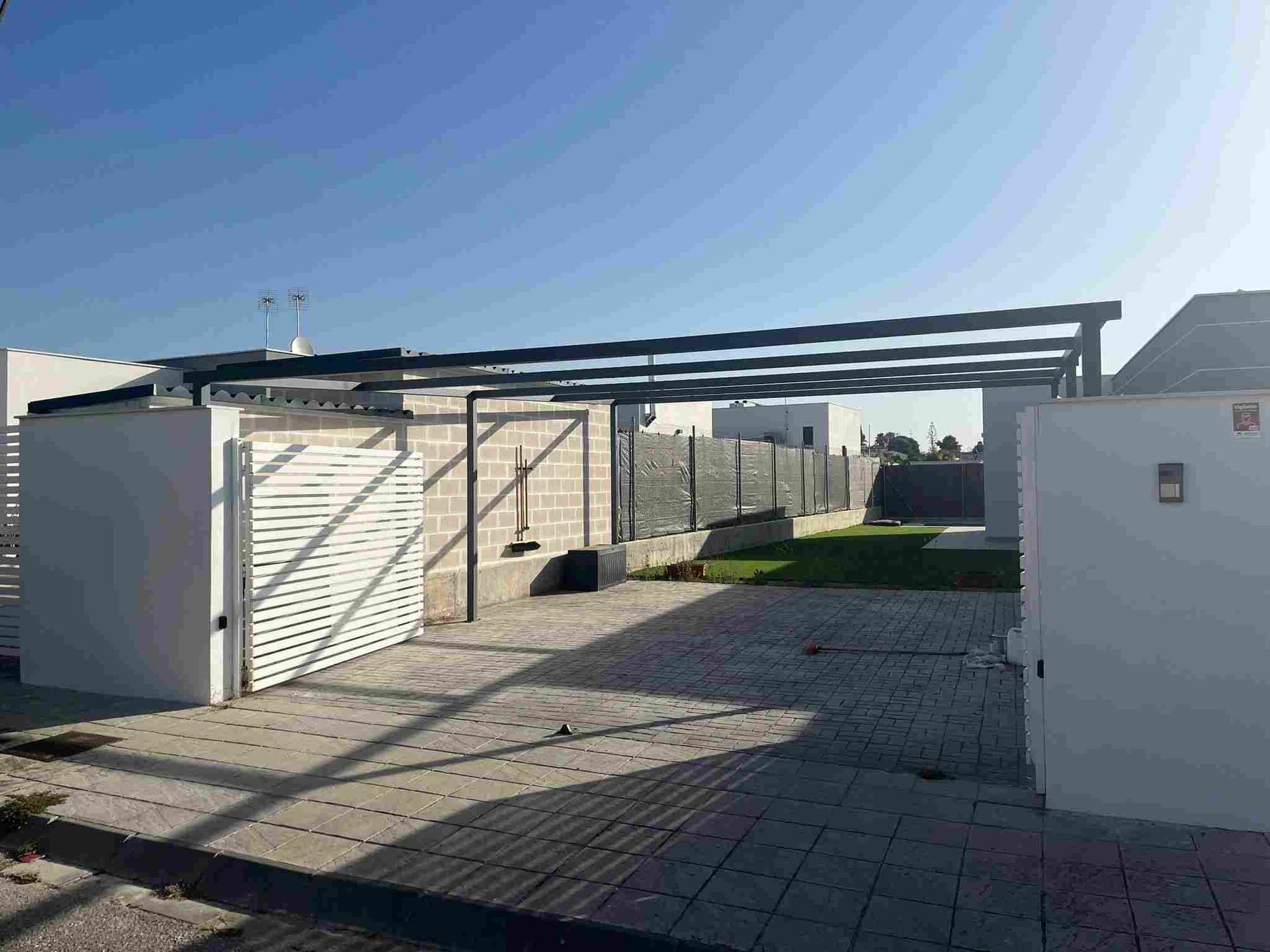 Cochera de metal sobre un camino de entrada pavimentado; cerca blanca y edificios al fondo bajo un cielo azul.