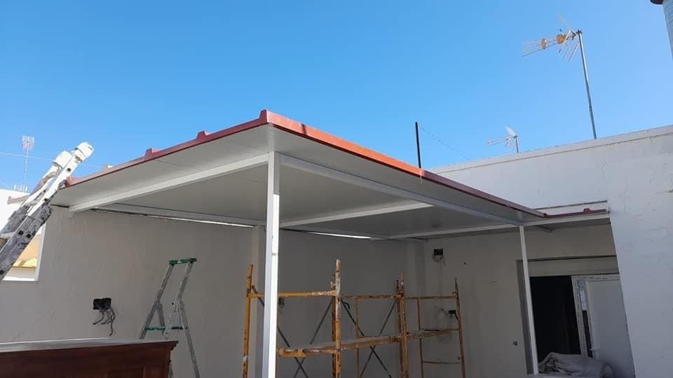 Un toldo exterior parcialmente construido con techo rojo, marco blanco y un cielo azul claro.
