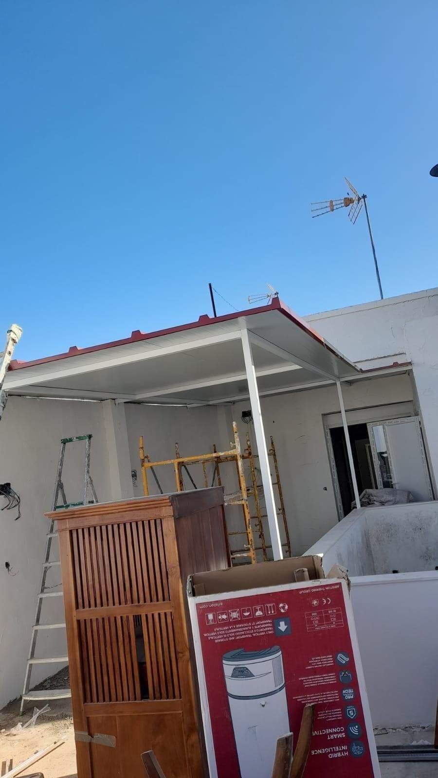 Techo de patio blanco adosado a un edificio blanco. Materiales de construcción y muebles abarrotan el primer plano. Cielo azul.