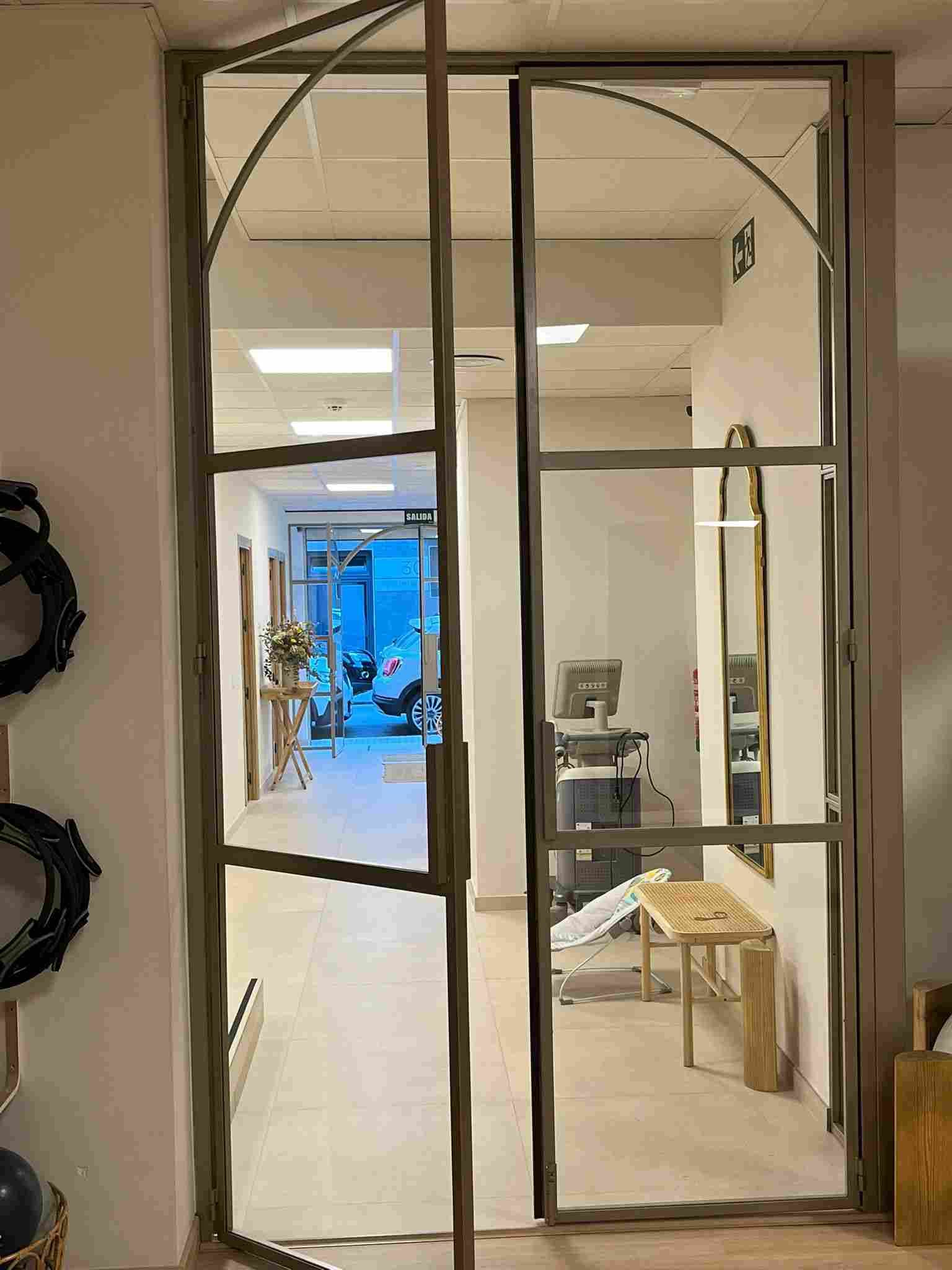 Puerta de vidrio con marco de metal, que conduce a una sala con equipo médico y mobiliario.