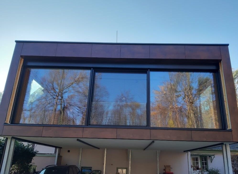 Modernes Gebäude mit großen Fenstern, in denen sich die Bäume spiegeln, unter einem Carport. Braune Fassade.