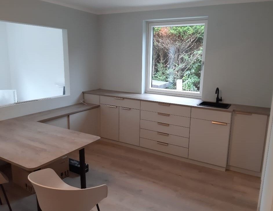 Eine kleine Küche mit hellen Schränken und einer Arbeitsplatte, einem Fenster mit Blick nach draußen und einem Tisch mit Stühlen.