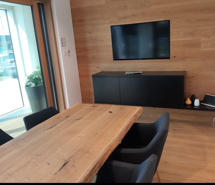 Esszimmer mit Holztisch, schwarzen Stühlen und einem an der Wand über einem schwarzen Schrank an einer holzgetäfelten Wand montierten Fernseher.