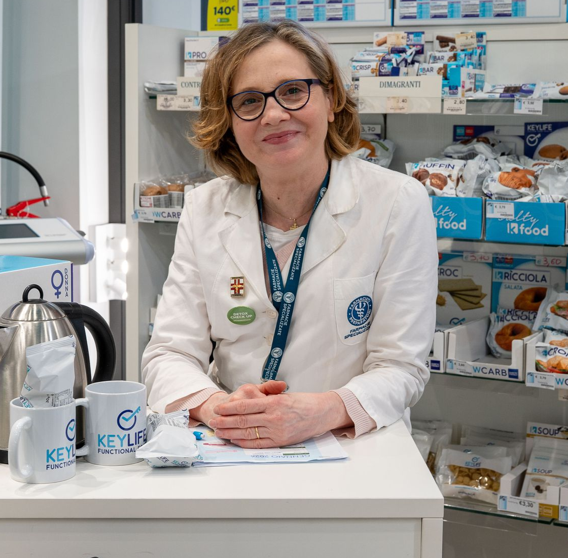 Farmacista in camice bianco che parla con un paziente, sullo sfondo sfocato gli scaffali della farmacia.