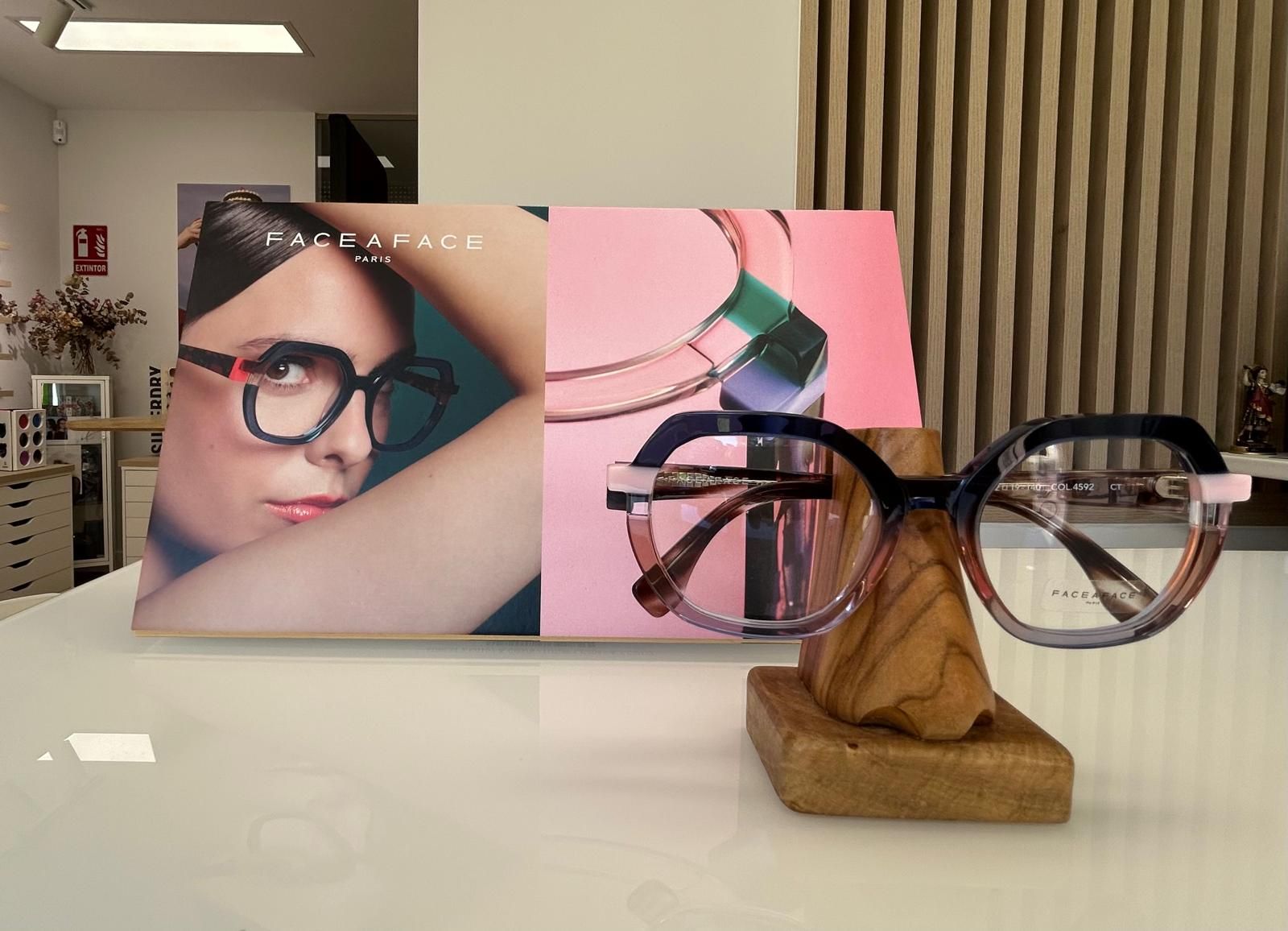 Gafas expuestas sobre un soporte de madera, con un póster de una mujer con gafas detrás.