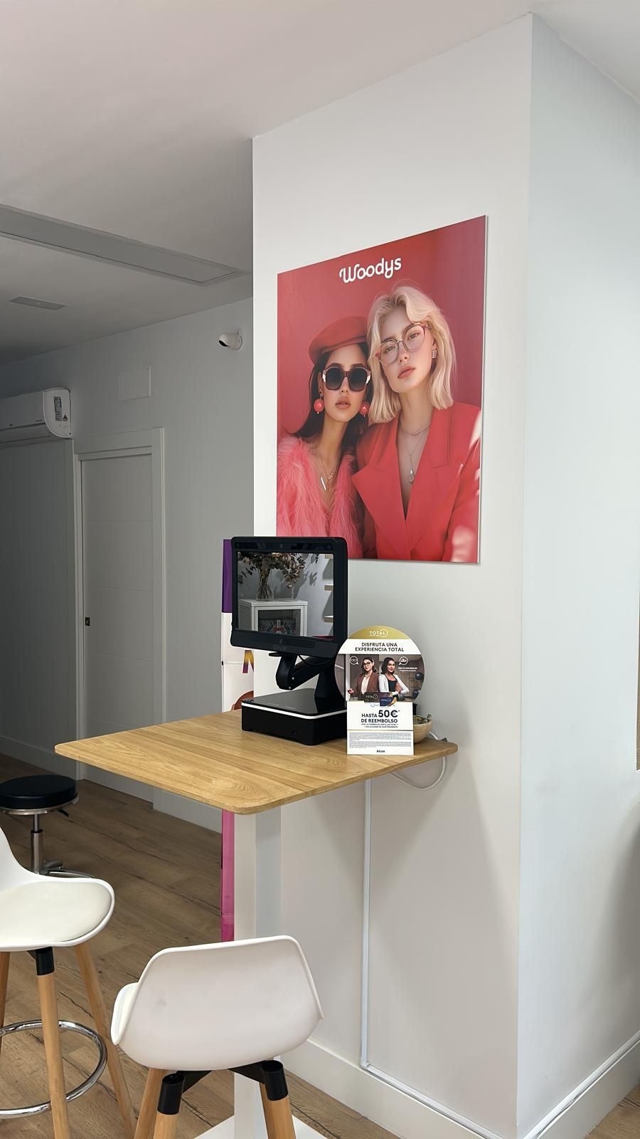 Rincón de cafetería pequeño con taburetes blancos, una mesa de madera fijada a la pared y un póster de moda rojo.