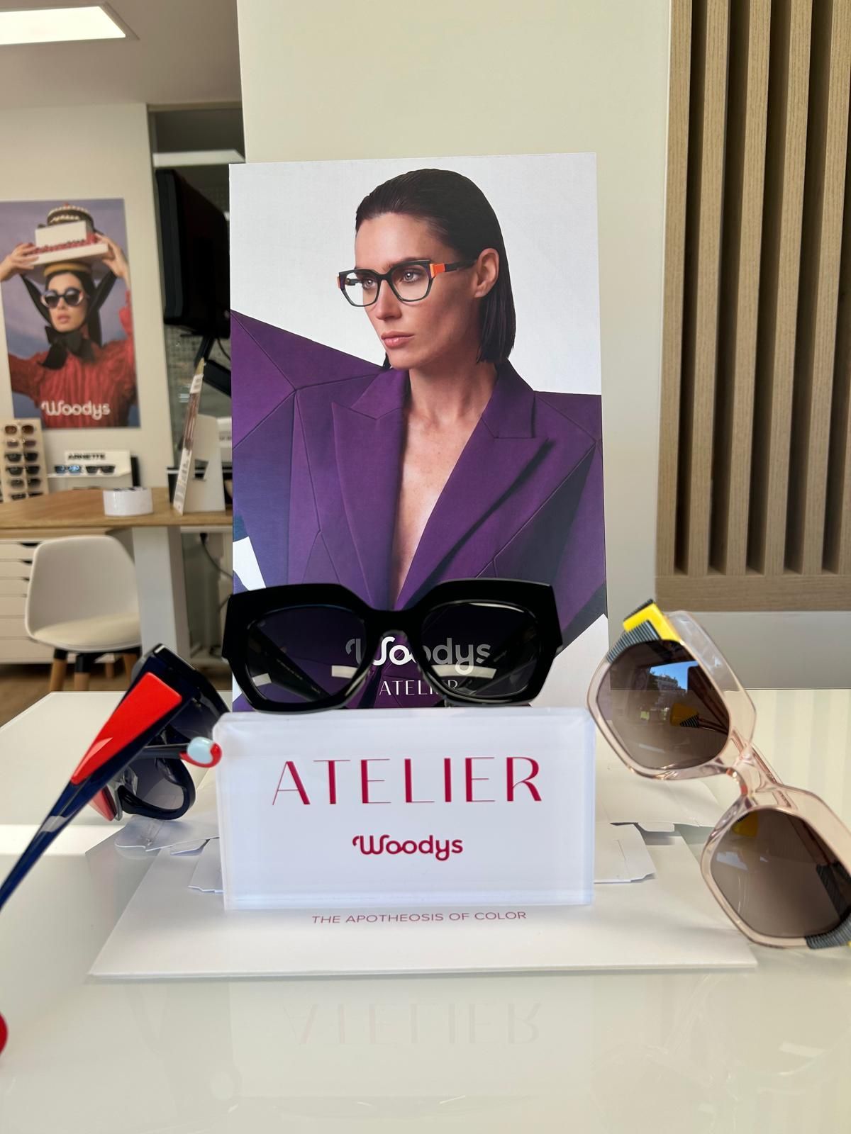 Gafas sobre una mesa blanca junto a un letrero que dice «ATELIER» y un póster de una modelo vestida de morado