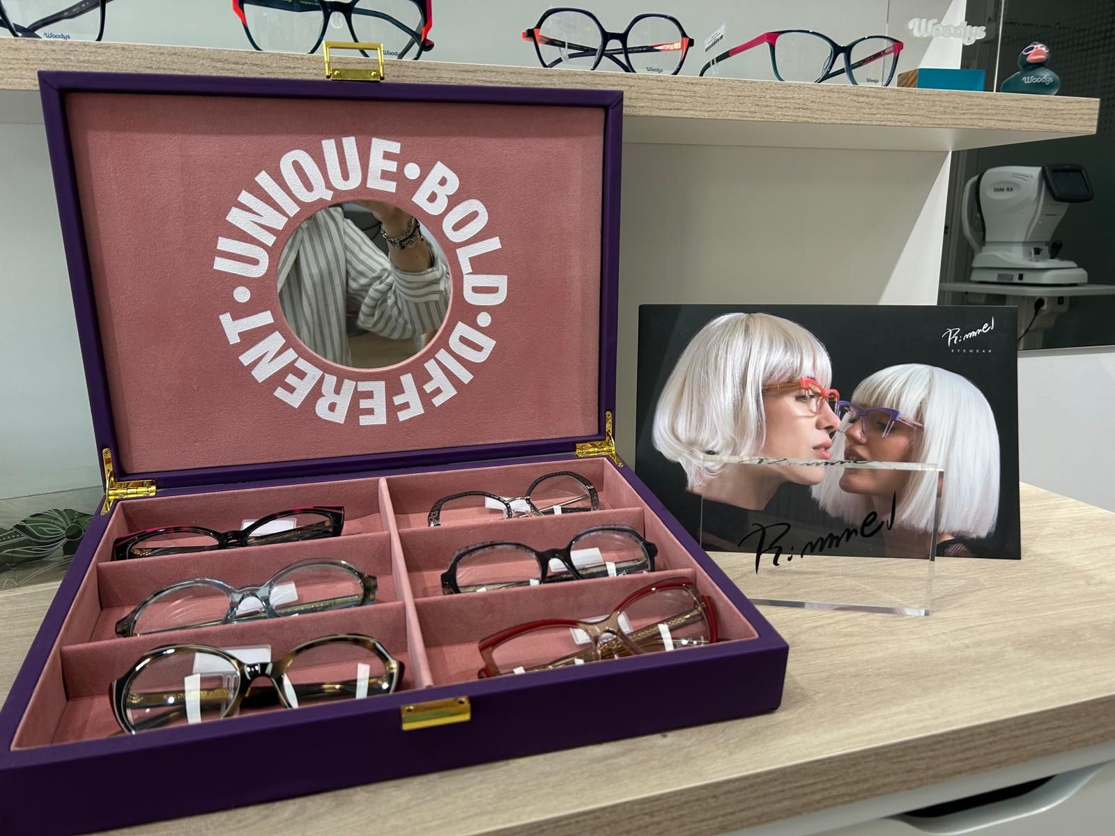 Estuche abierto de gafas morado con varias monturas junto a un espejo que muestra a dos personas de pelo blanco tocándose la frente