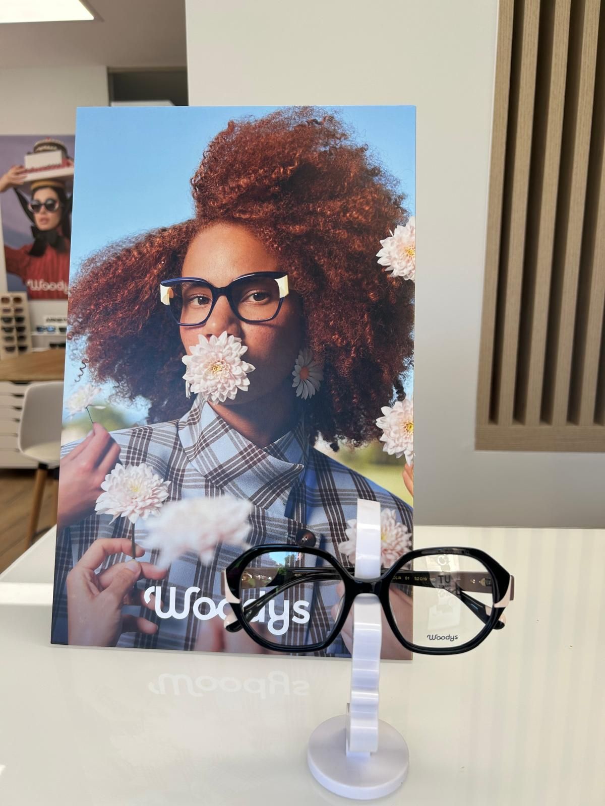 Gafas negras sobre un soporte transparente, delante de un cartel con una persona y flores.