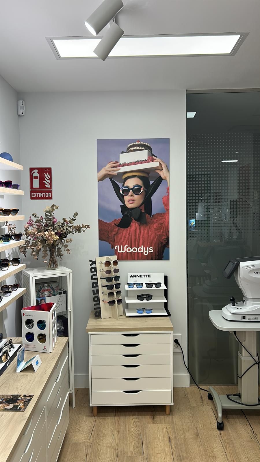 Expositor de una óptica con gafas, armarios blancos, un póster y un lavabo en un probador bien iluminado