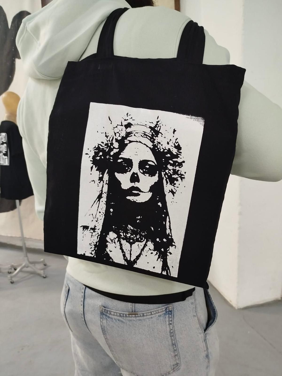 Bolso tote negro con estampado facial monocromático, usado sobre una camisa blanca y jeans en un ambiente interior.
