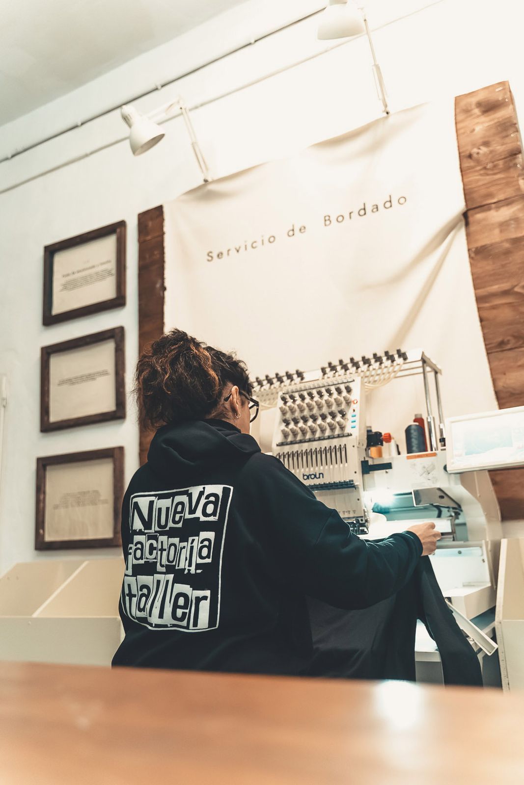 Persona sentada en escritorio en un estudio, trabajando con materiales de costura, con una obra en la pared de fondo.