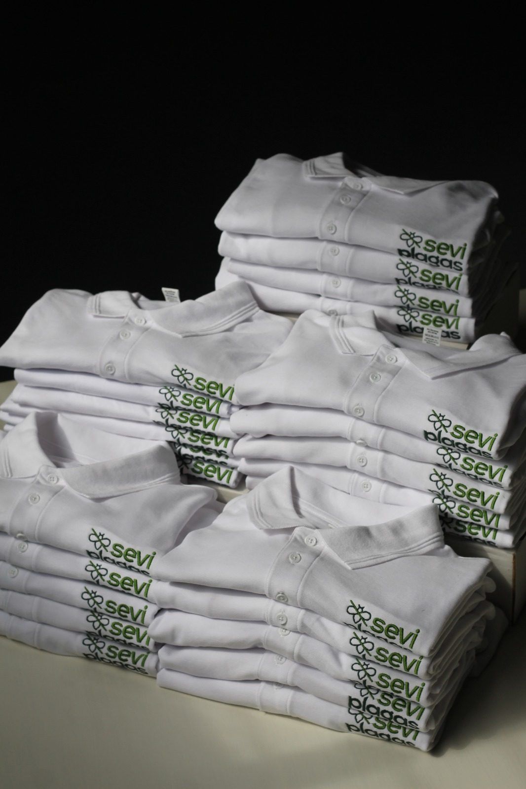 Camisetas blancas dobladas apiladas con etiquetas verdes impresas sobre un fondo oscuro.