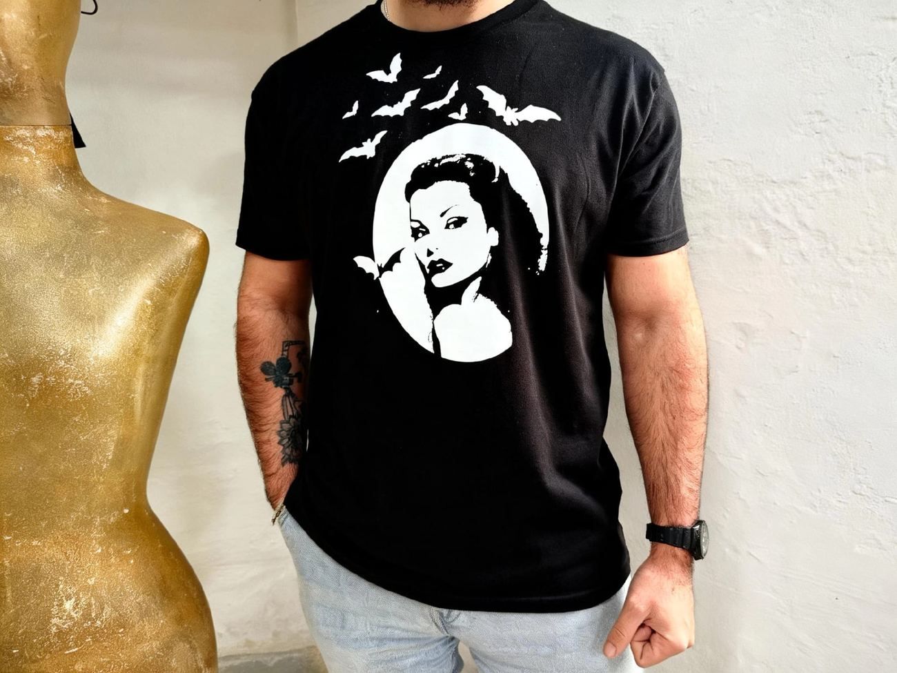 Persona con camiseta negra con estampado blanco, de pie junto a un torso de maniquí dorado contra una pared blanca.