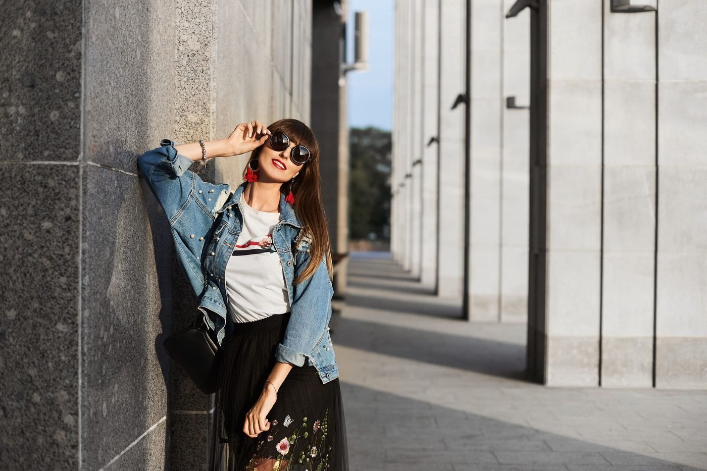 Mujer con chaqueta vaquera apoyada contra un muro de piedra bajo la luz del sol, con gafas de sol y sonriendo.