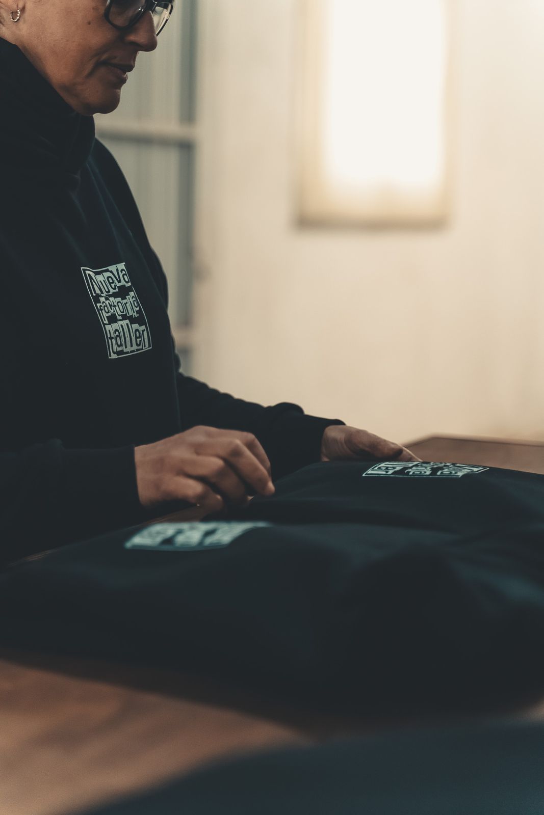 Una persona con una camisa negra manipula una tela doblada sobre una mesa en una habitación con iluminación tenue.