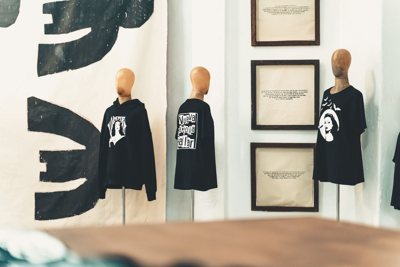Tres camisas negras sobre maniquíes en una galería junto a obras de arte enmarcadas y certificados.