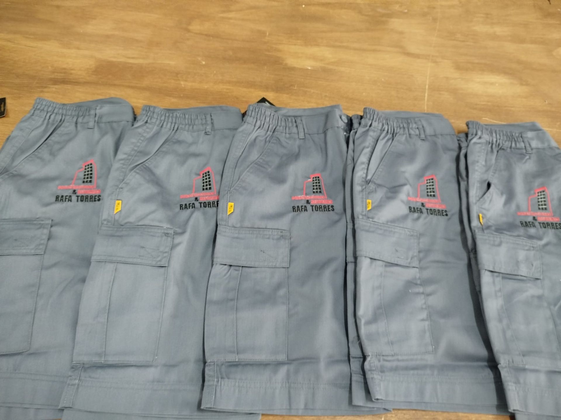 Cinco pantalones de trabajo grises, sobre un suelo de madera, con parches reflectantes con el logotipo en los bolsillos.