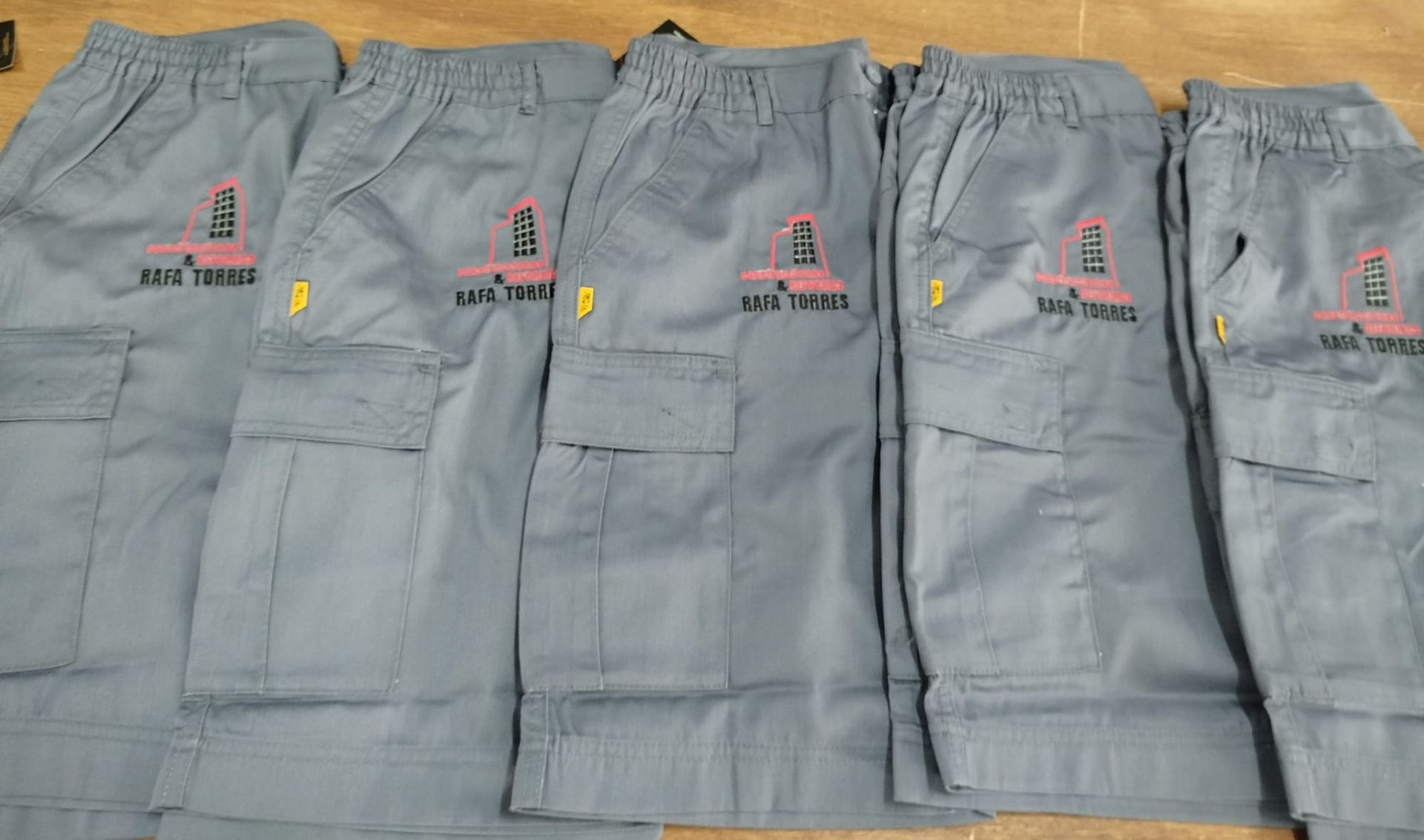 Fila de cinco pantalones de trabajo grises con logotipos bordados colocados uno al lado del otro
