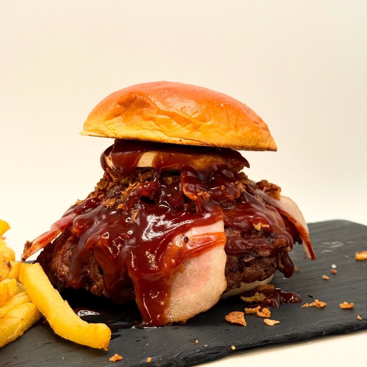 Una gran hamburguesa con queso, tocino y salsa, junto a una pila de papas fritas, en un plato.