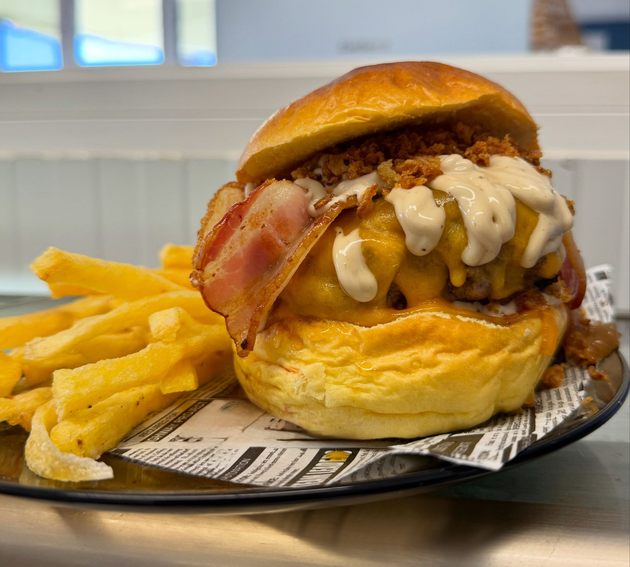 Una gran hamburguesa con queso, tocino y salsa, junto a una pila de papas fritas, en un plato.