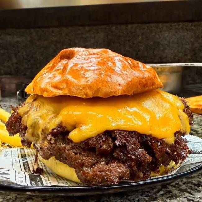 Una gran hamburguesa con queso, tocino y salsa, junto a una pila de papas fritas, en un plato.