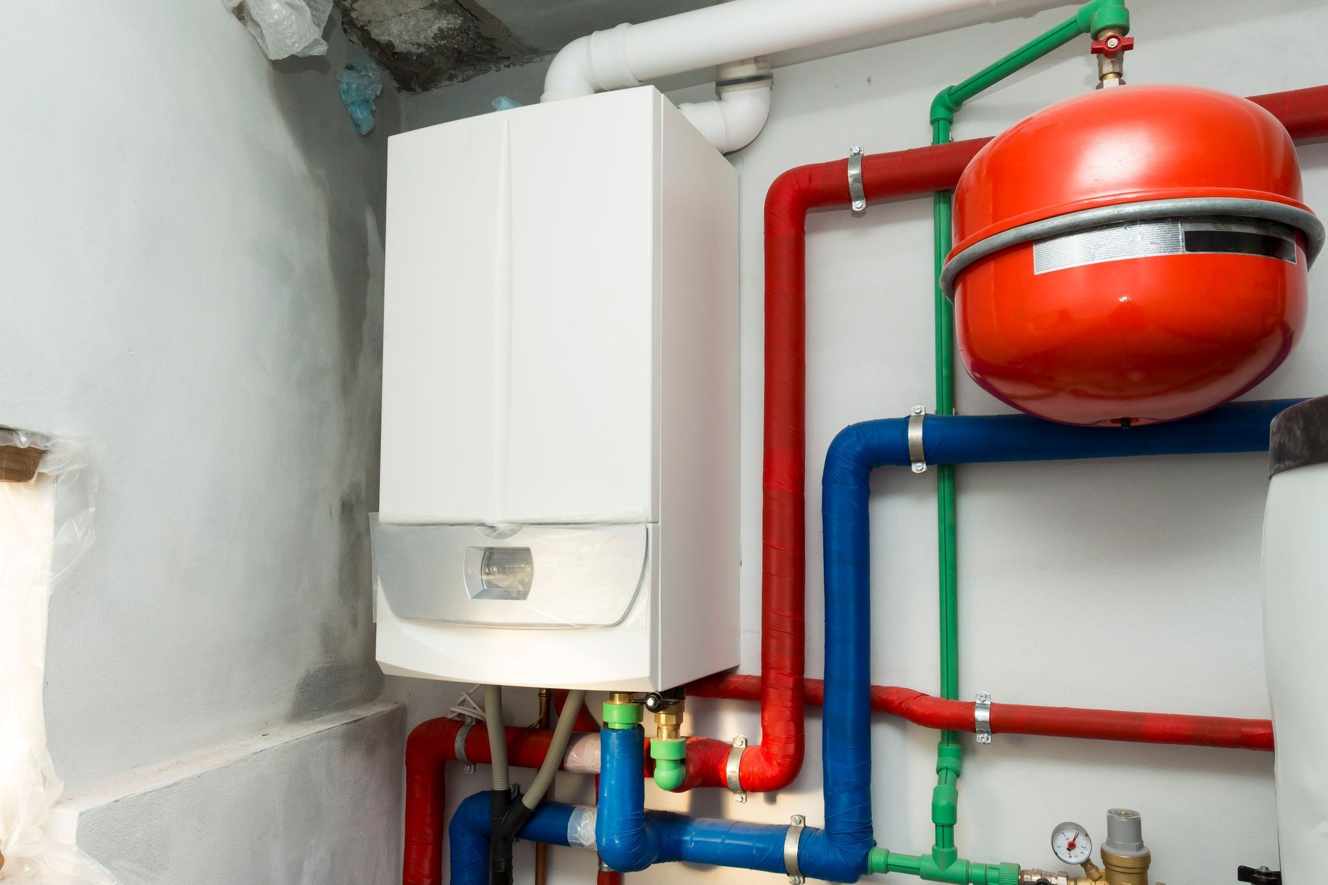Chaufferie avec chauffe-eau blanc, tuyaux rouges et bleus, et vase d'expansion rouge fixé au mur.