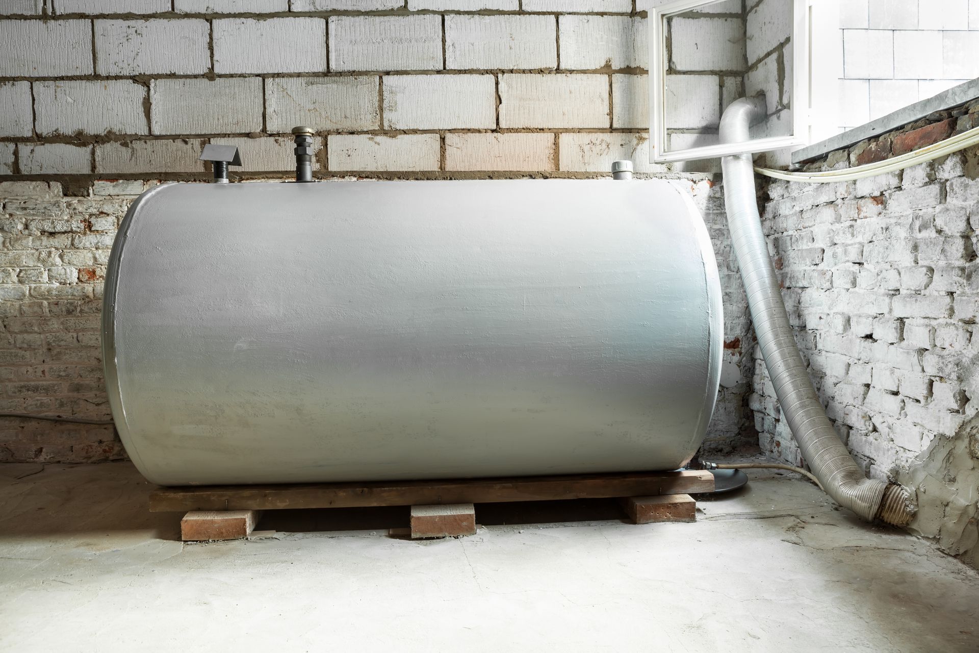 Grand réservoir cylindrique gris posé sur des palettes en bois dans un sous-sol aux murs de briques.