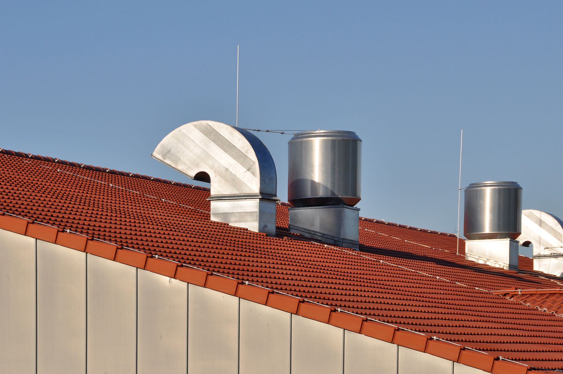 Toit avec des tuiles rouges, des aérations argentées et une antenne parabolique blanche se détachant sur un ciel bleu.