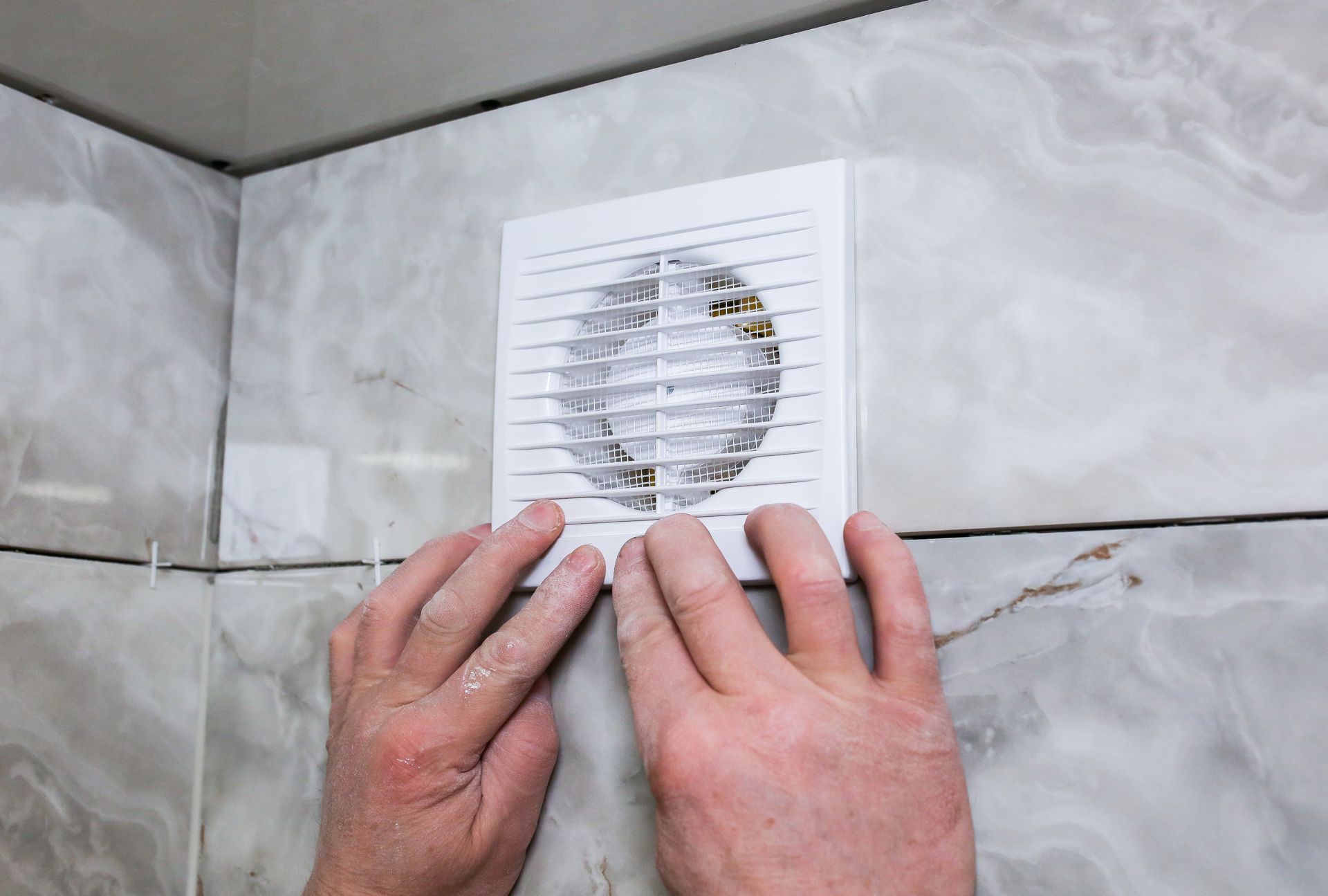 Mains installant une grille d'aération blanche pour salle de bain sur un mur carrelé.