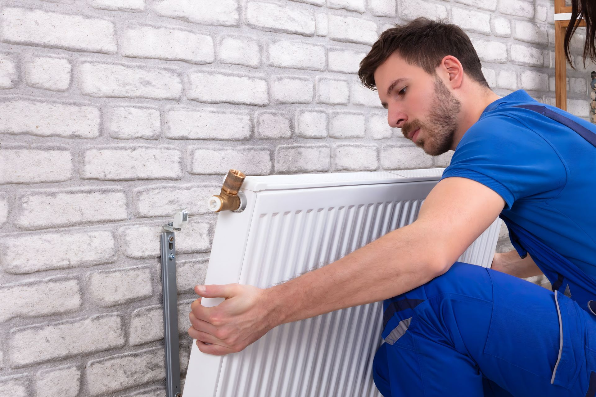 Un homme en tenue de travail bleue installe un radiateur blanc contre un mur de briques.