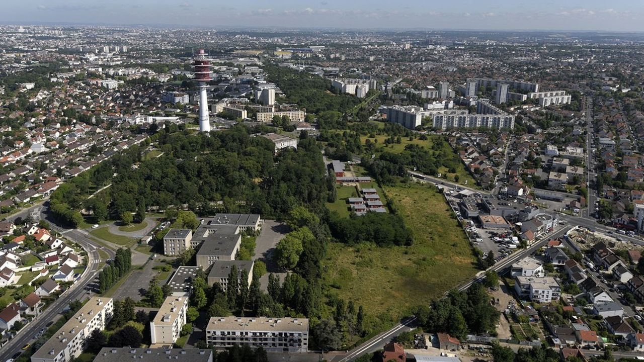 Vue aérienne d'une ville aux quartiers denses, avec un grand parc verdoyant et une haute tour blanche près du centre.