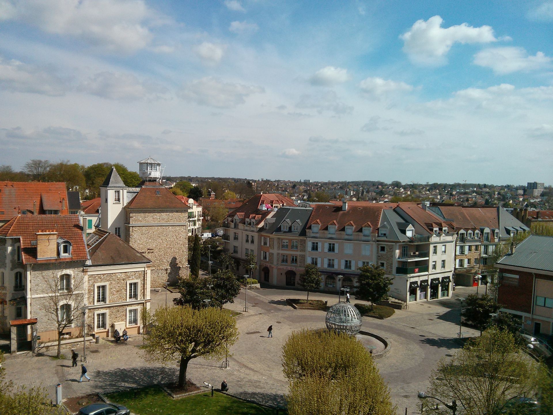 Vue aérienne d'une place de ville européenne avec des bâtiments historiques, des toits rouges et des arbres sous un ciel partiellement nuageux.