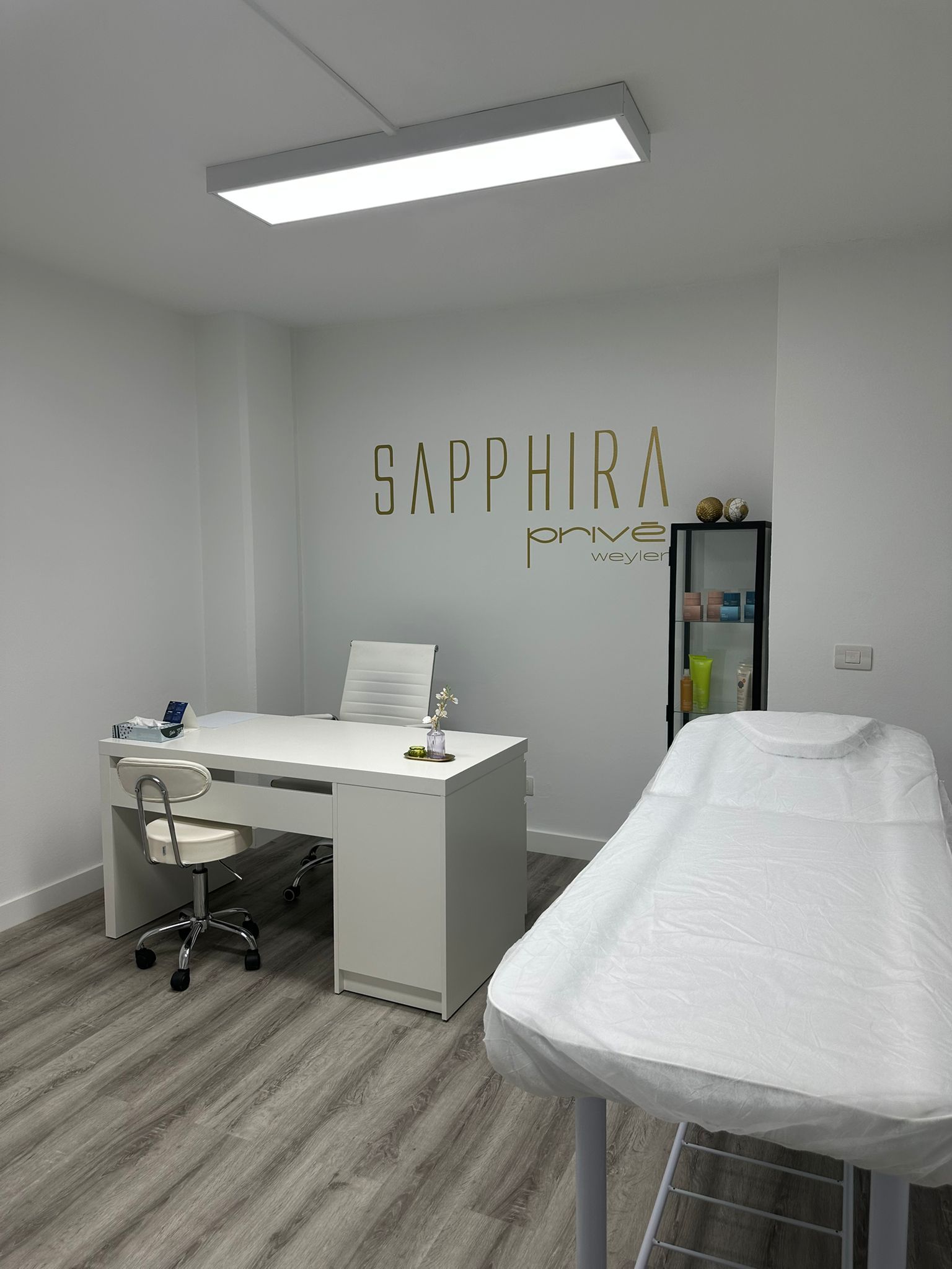 Sala de tratamientos de spa con paredes blancas, camilla de masaje y productos para el cuidado de la piel. Logotipo dorado en la pared.