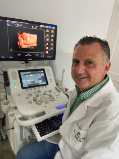 Un médico sonriente junto a una máquina de ultrasonido que muestra una exploración en 3D en una clínica.