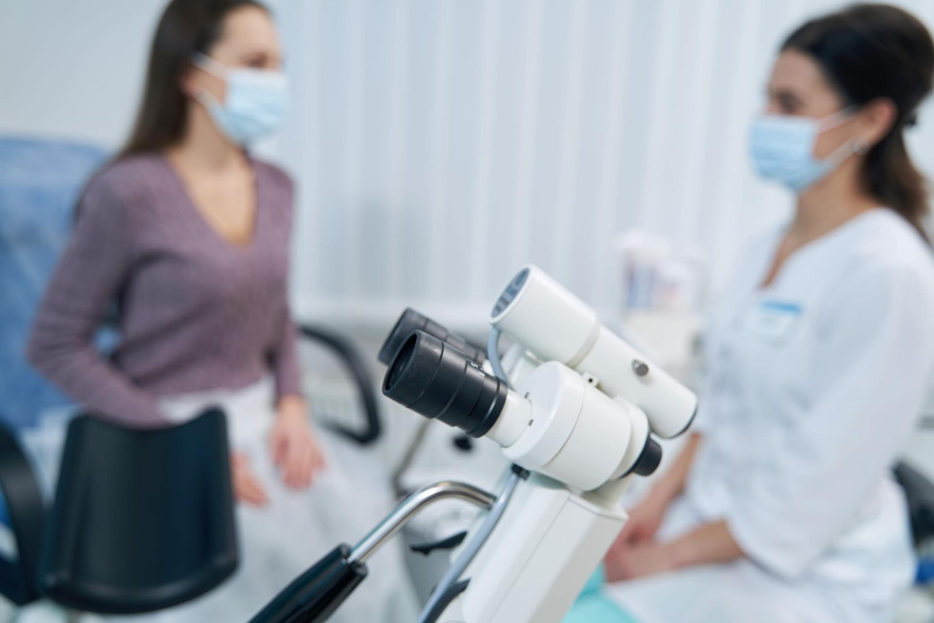 Dos mujeres enmascaradas en una consulta médica con un microscopio en primer plano.