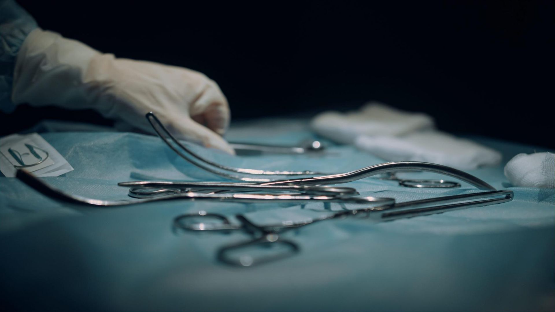 Mano enguantada sosteniendo pinzas quirúrgicas sobre un paño estéril azul con instrumentos médicos.