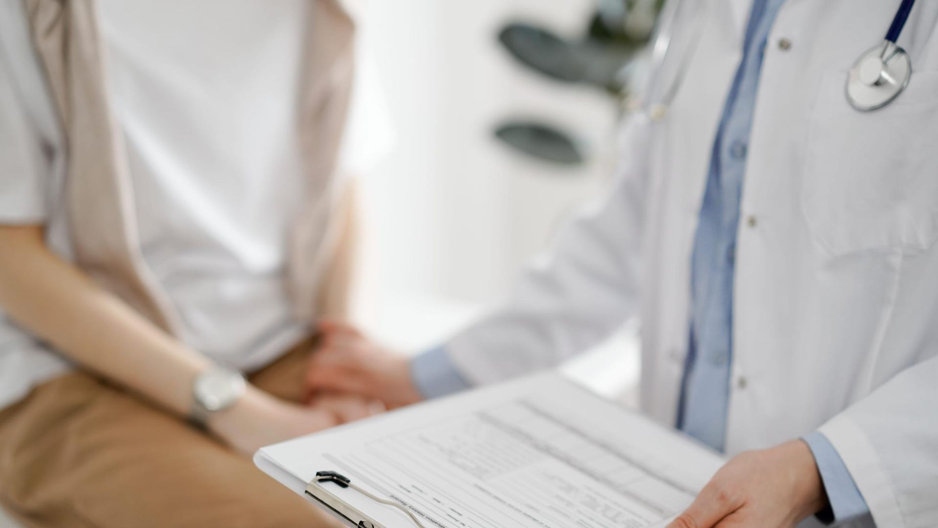 Un médico sostiene un portapapeles mientras habla con un paciente sentado en un consultorio médico.