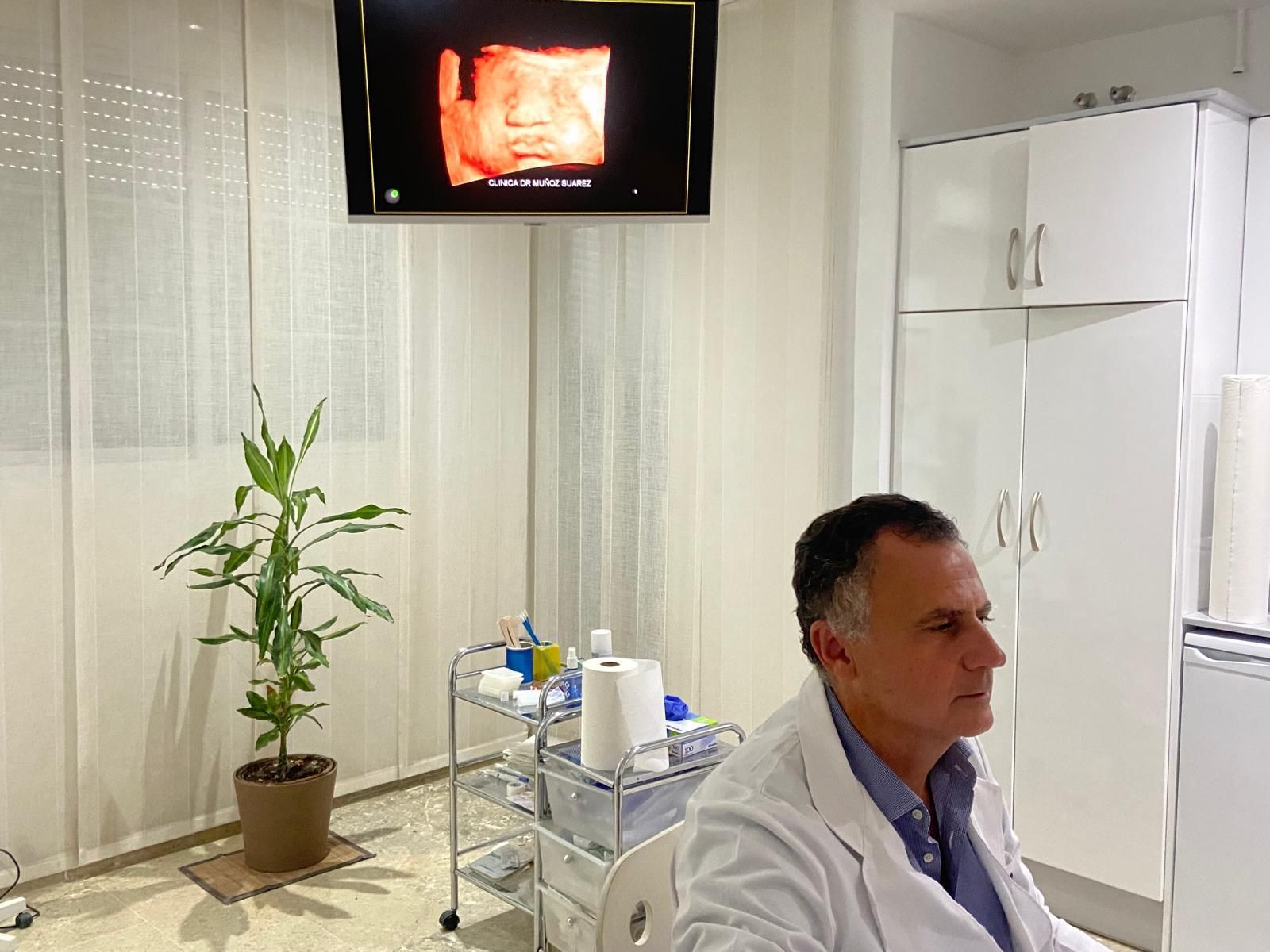 Médico con bata blanca sentado en una sala de consulta con equipo médico y un monitor montado en la pared