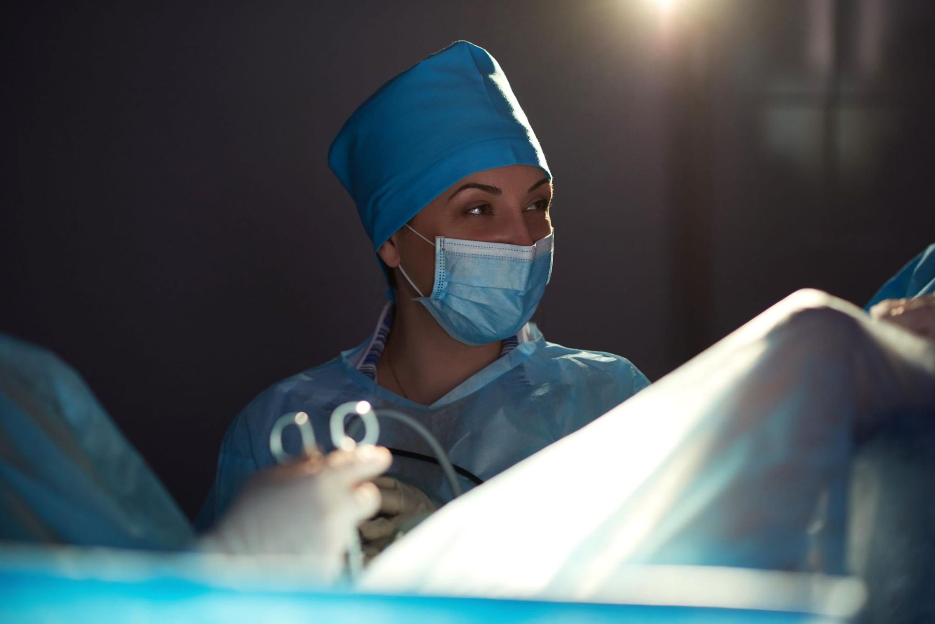 Cirujano con bata azul y mascarilla en un quirófano, concentrado en un paciente bajo luces quirúrgicas brillantes.