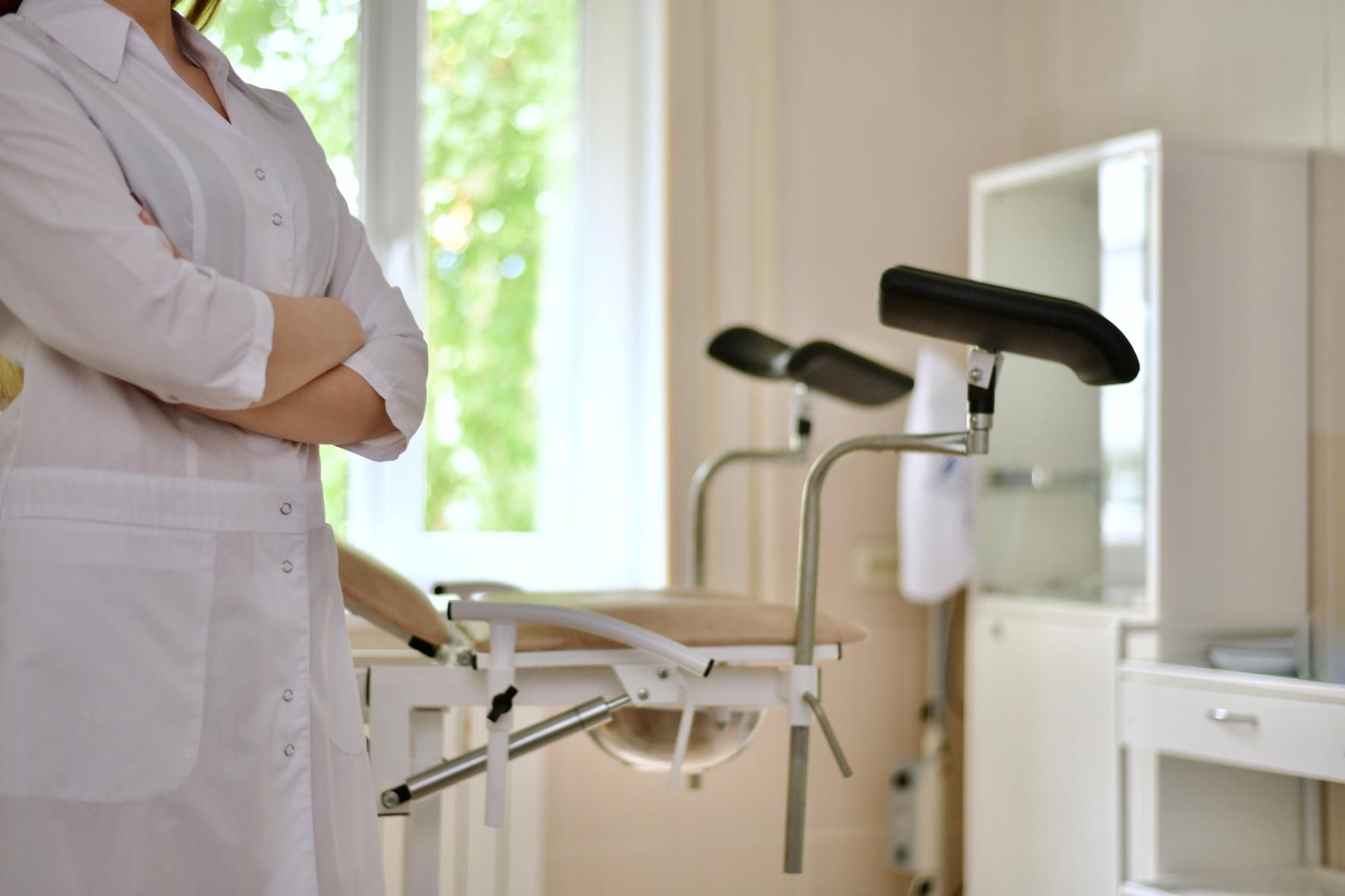 Sala de exploración médica con un médico vestido de blanco de pie junto a una silla de exploración ginecológica.
