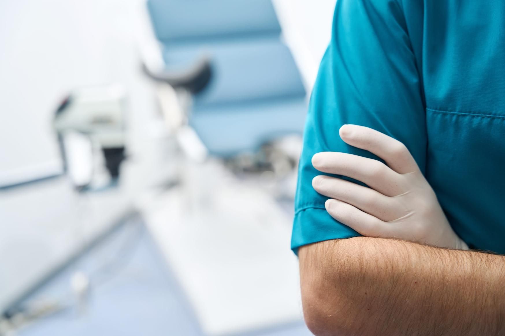 Persona con camisa azul sosteniendo un brazo, con un fondo borroso de un consultorio médico.