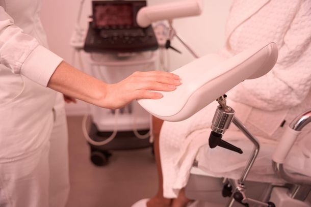 Ajustando manualmente una silla clínica blanca junto a una camilla de exploración ginecológica.