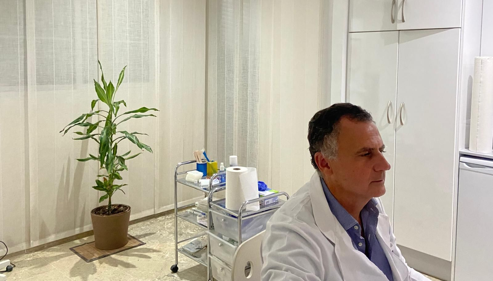 Hombre con bata blanca sentado en un luminoso consultorio médico junto a un carrito con plantas y equipos.