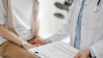 Un médico sostiene un portapapeles mientras habla con un paciente sentado en un consultorio médico.
