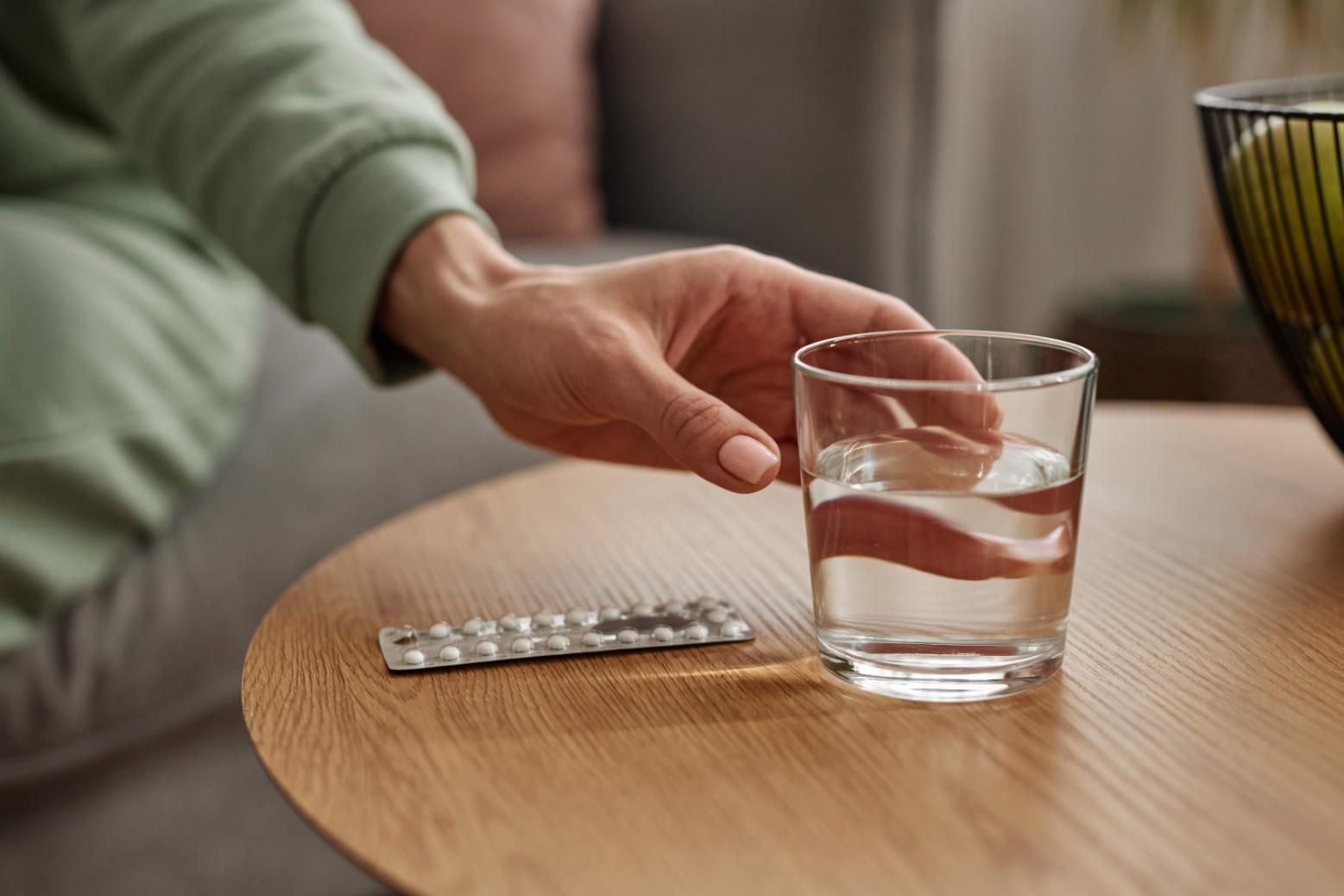 Una mano se extiende hacia un vaso de agua sobre una mesa de madera, junto a un blíster de pastillas.