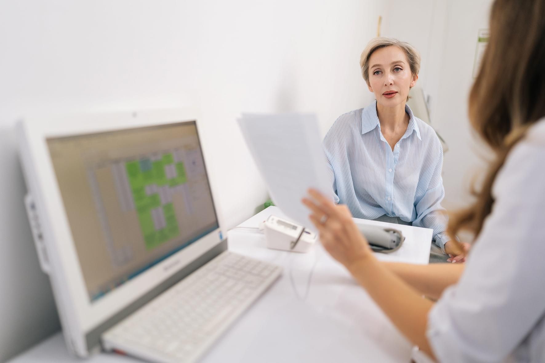 Dos personas en una consulta luminosa discutiendo documentos frente a frente, con una computadora portátil abierta.