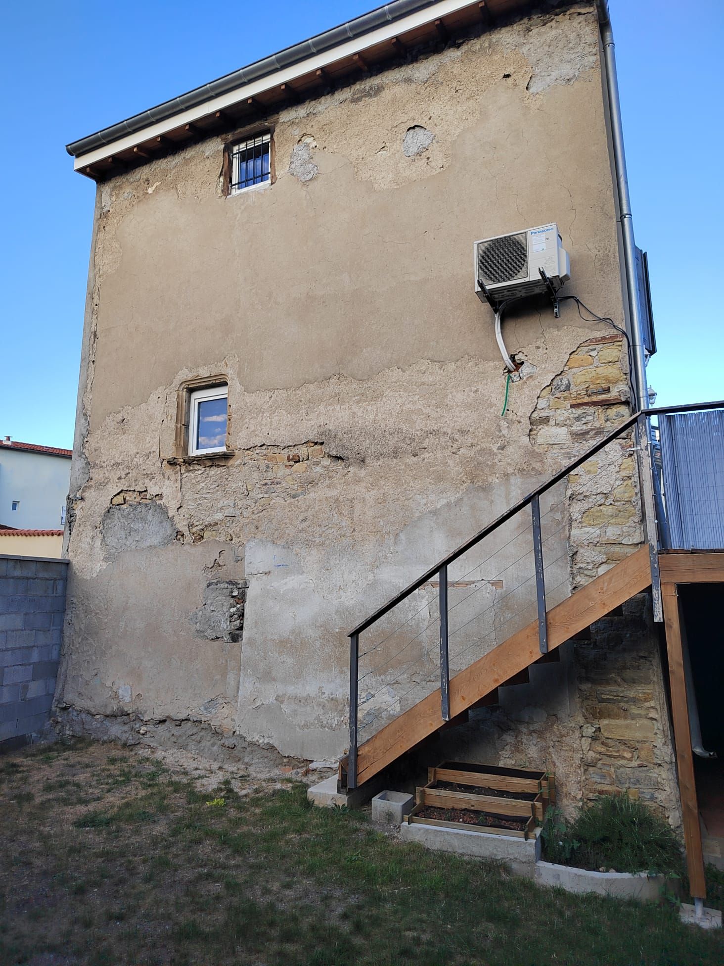 Bâtiment en stuc beige avec climatisation, deux fenêtres et escalier en bois.