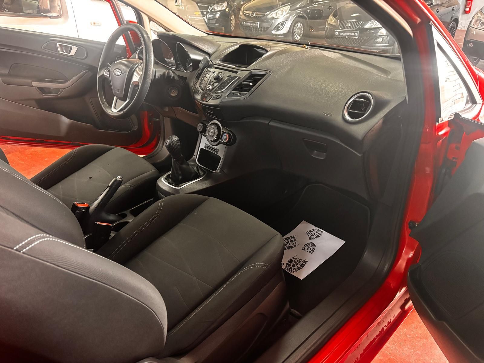 El interior de un coche rojo cuenta con asientos de tela negra, transmisión manual y salpicadero negro.