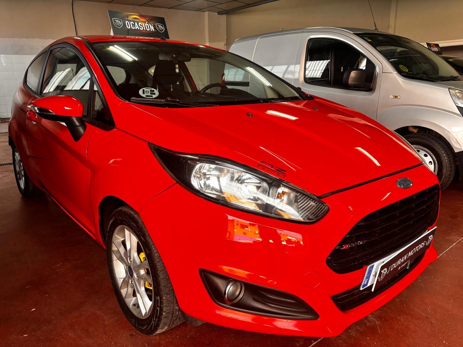 Un Ford Fiesta hatchback de color rojo brillante estacionado dentro de un garaje junto a una furgoneta comercial plateada.